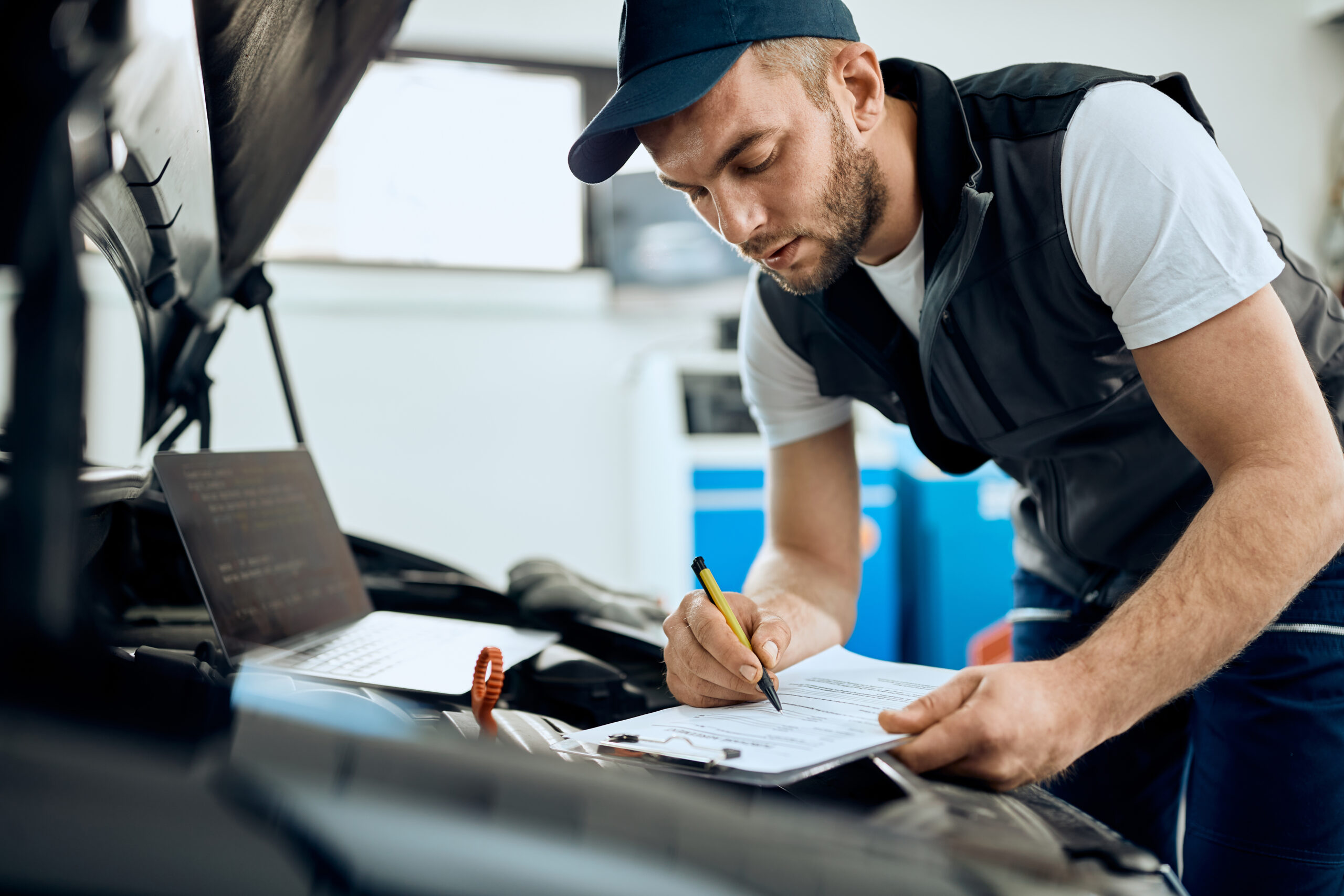 Young repairman going through checklist while examining car engine in a workshop.